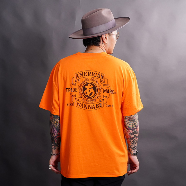 THE AMERICAN WANNABE/アメリカンワナビー「TRADE MARK S/S TEE / ORANGE」プリントS/S Tシャツ