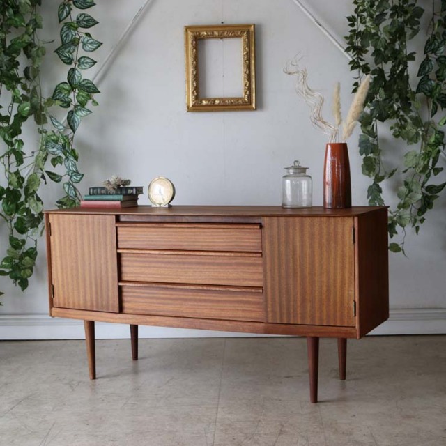 チーク材の木目が美しいサイズが小さめの脚付きサイドボート、両側が開き戸で真ん中は３段の引き出し